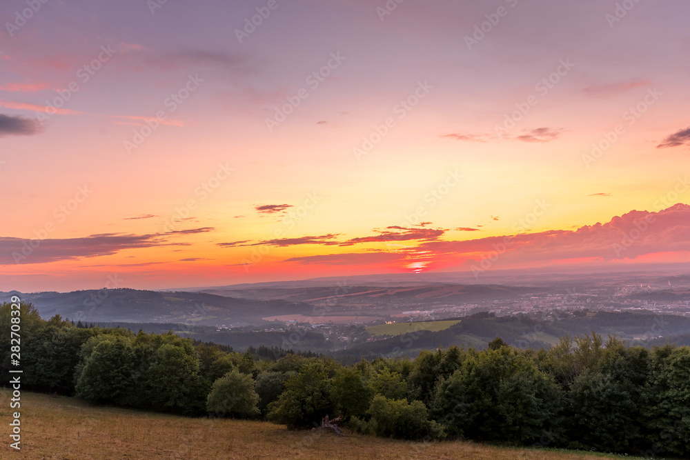 Sunset with view on landscape with fully colored clouds orange and purple and sun behind it and city Valasske Mezirici captured during summer late time.