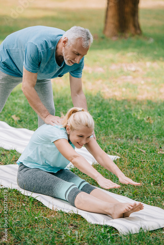 handsome mature man helping smiling woman practicing stretching yoga pose on lawn