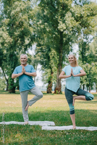 mature man and woman meditating in tree poses while standing in yoga mats in park