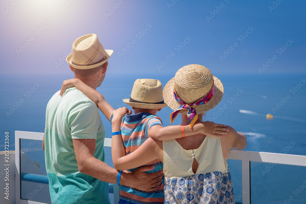 Family of three people on hotel balcony against sea in summer enjoying their vacation. Back view.