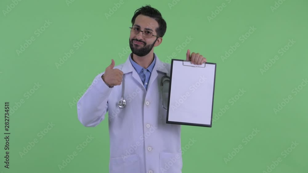 Happy young bearded Persian man doctor showing clipboard and giving thumbs up Happy young bearded Persian man doctor showing clipboard and giving thumbs up