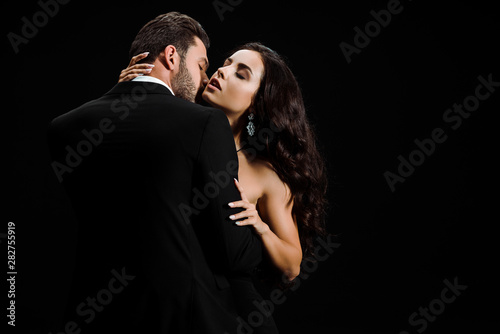passionate man in suit kissing attractive woman isolated on black