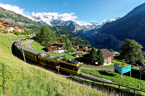 Berner Oberland, Switzerland - july 12: Tourist train traveling on the railway thru Wengen village by green grassy hillside with Jungfrau mountain & Lauterbrunnen Valley in background on a summer day 