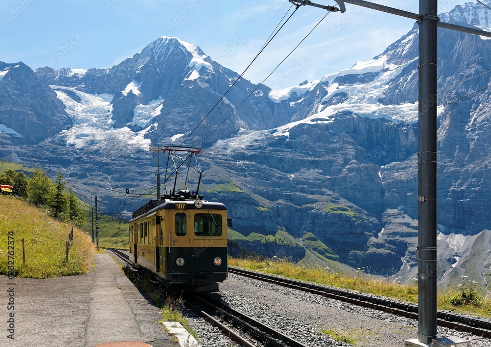 A cog wheel train traveling on the mountain Railway from Wengen to Kleine Scheidegg station with majestic Swiss Alps ( Eiger, Monch, Jungfrau ) in background, in Bernese Highlands, Switzerland, Europe
