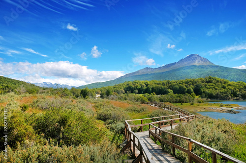 Panoramic view of Tierra del Fuego National Park, showing mountains surrounded by green vegetation and water, against a blue sky.