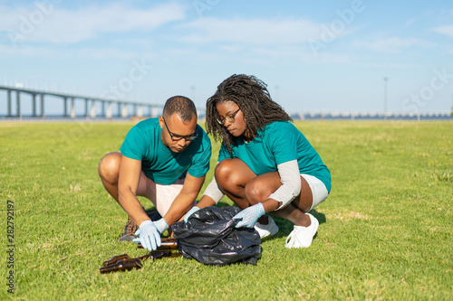 Volunteers working on lawn and collecting empty bottles into plastic bag. Black woman and Latin man wearing uniforms and protective gloves cleaning lawn from garbage. Litter collection concept © Mangostar