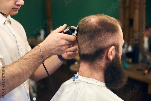 Perfect trimming. Back side close-up view of young bearded man getting haircut by masterful barber with electric razor while sitting in hairdresser chair. Contemporary service in barbershops.