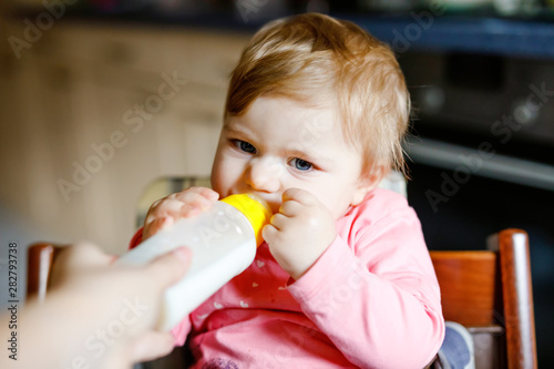Cute adorable baby girl holding nursing bottle and drinking formula milk. First food for babies. New born child  sitting in chair of domestic kitchen. Healthy babies and bottle-feeding concept