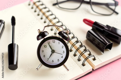 Black glasses, alarm clock, paper notepad, mascara and red pomade on pink background composition.