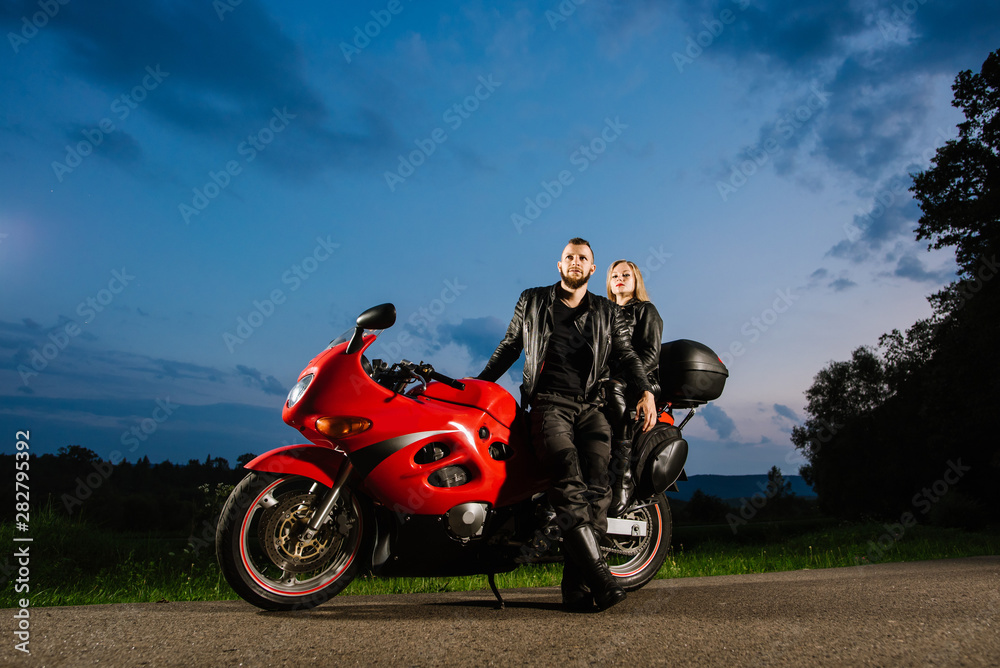 Obraz premium Cool biker couple in leather jackets sitting on red sports bike outdoors