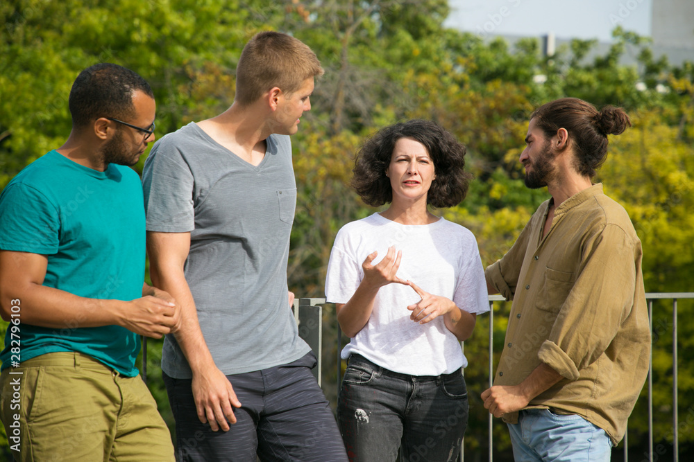 Focused pensive female group leader giving instruction. Middle aged woman standing outdoors, speaking, count on fingers, team of young men listening to her. Group leader concept Focused pensive female group leader giving instruction. Middle aged woman standing outdoors, speaking, count on fingers, team of young men listening to her. Group leader concept