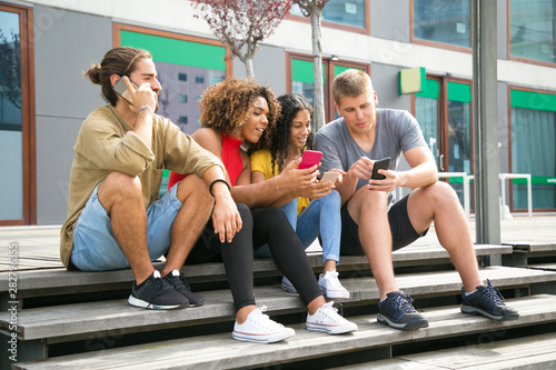 Excited group of friends using mobile phone outside. Mix raced girls and guy showing smartphone screens to each other, young man talking on cell. Digital gadgets concept