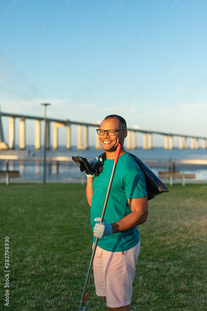 Happy male volunteer posing in city park. Outdoor portrait of Latin man standing on grass, holding rake and plastic bag, smiling at camera. Happy volunteer concept