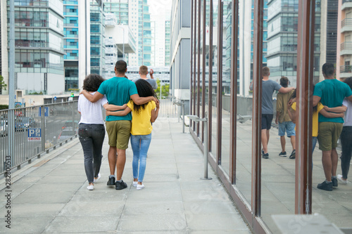 Team of male and female friends enjoying outdoor walk. Back view of mix raced people walking down city street, hugging each other and talking. Polygamy concept photo