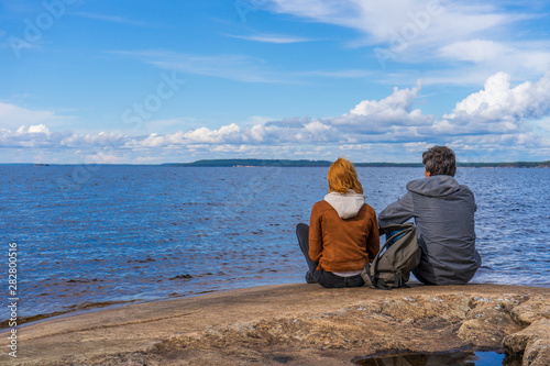 Tourists man and woman sitting on northern lake shore in summer day. People relaxing and admiring beautiful landscape. Travelling and discovering distant places of Earth. Onega lake, Karelia, Russia