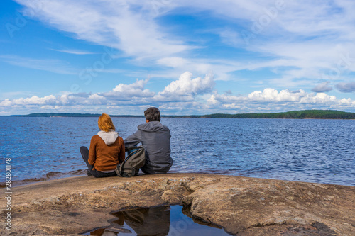 Tourists man and woman sitting on northern lake shore in summer day. People relaxing and admiring beautiful landscape. Travelling and discovering distant places of Earth. Onega lake, Karelia, Russia