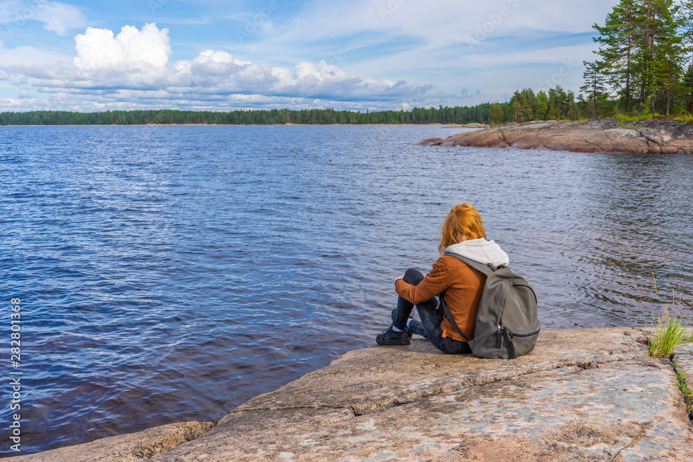 Young tourist lady sitting on northern lake shore in summer day. Teenage girl relaxing and admiring beautiful landscape. Travelling and discovering distant places of Earth. Onega lake, Karelia, Russia