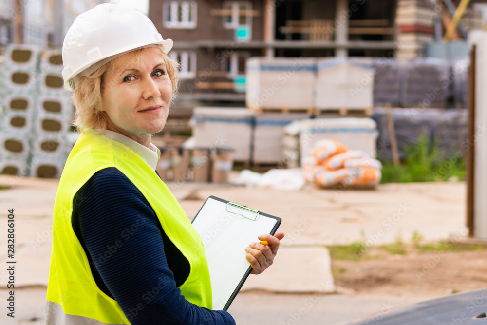 Builder woman on construction site in green vest and white helmet with tablet. A middle-aged woman looks at the camera. She writes in a notebook. Builder woman on construction site in green vest and white helmet with tablet. A middle-aged woman looks at the camera. She writes in a notebook.
