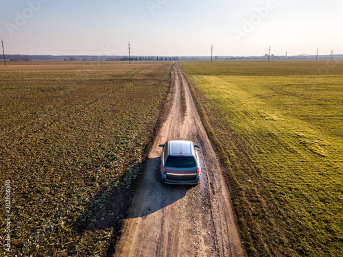 Aerial view of car driving by straight ground road through green fields on sunny blue sky copy space background. Drone photography.