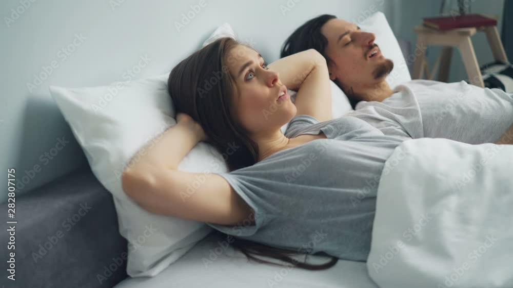 Irritated woman is covering head with pillow when boyfriend is snoring in sleep in bed disturbing her relaxation. Healthcare, problems and family concept. Irritated woman is covering head with pillow when boyfriend is snoring in sleep in bed disturbing her relaxation. Healthcare, problems and family concept.