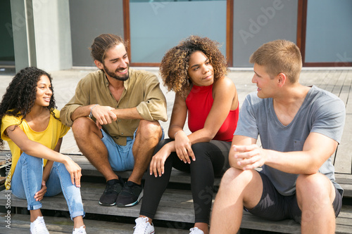 Happy friends meeting outdoors and discussing news. Multiethnic group of young people sitting on outdoor staircase, talking, listening, laughing. Close friends meeting concept