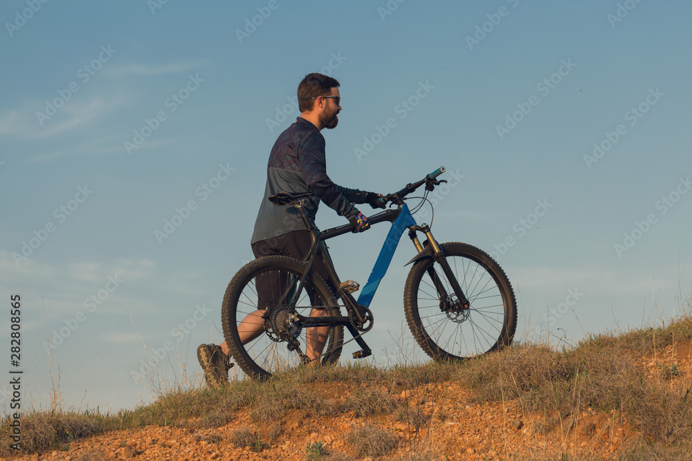 Cyclist in shorts and jersey on a modern carbon hardtail bike with an air suspension fork rides off-road on the orange-red hills at sunset evening in summer	