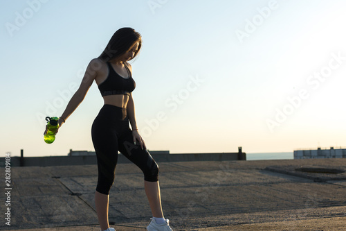 Slim athletic girl takes a break between classes and drinks water from a bottle, urban background. Beautiful sky at sunset.