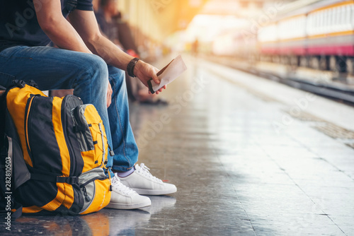 Cheap Travel and Lifestyle concept. Holiday Time, Young Traveler man wearing sneaker and sitting at train station. Asian Backpacker waiting train alone and plan trip in summer time with smart phone .