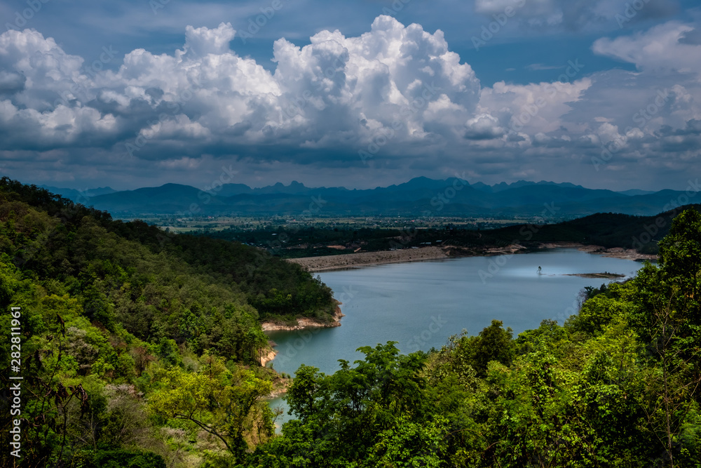 View of the reservoir surrounded by mountains and green forest from the mountain top against cloudy sky with white fluffy cumulus clouds.