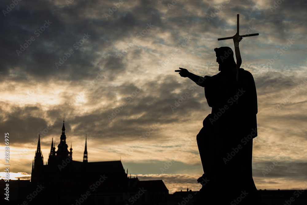Naklejka premium Silhouette of St. John the Baptist statue on Charles Bridge in Prague with St. Vitus Cathedral in background at sunset.