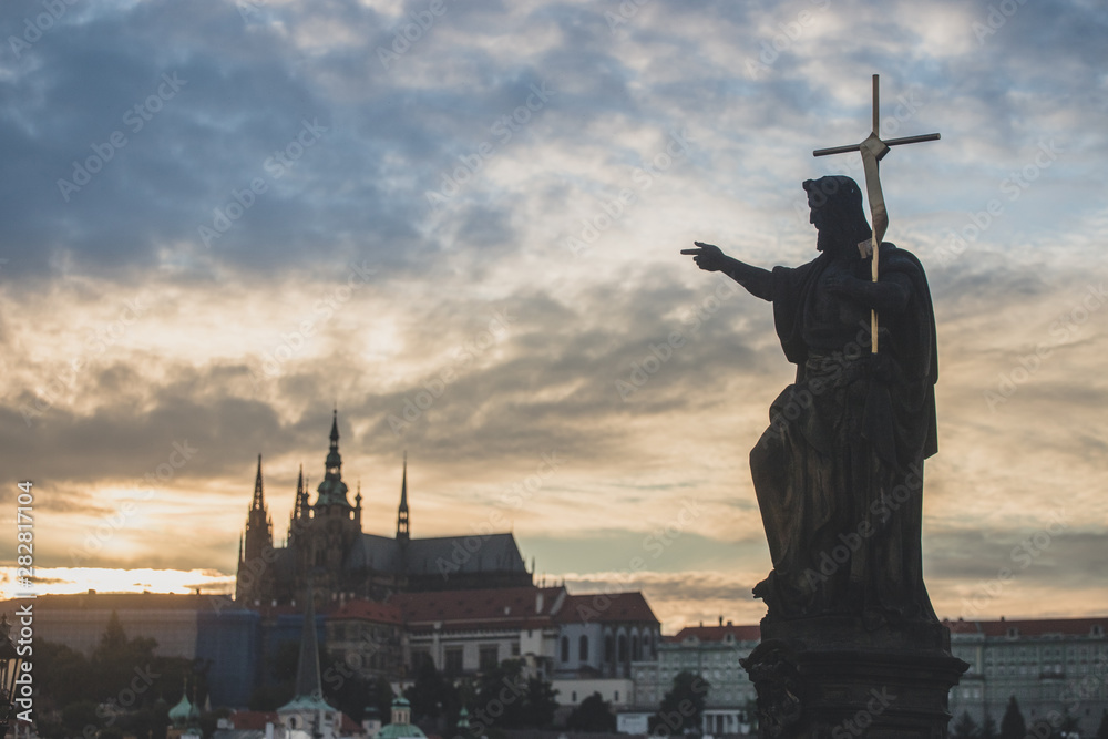 Naklejka premium St. John the Baptist statue on Charles Bridge in Prague with St. Vitus Cathedral in background at sunset.