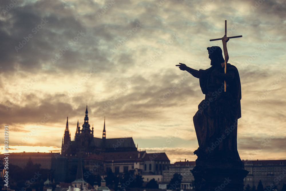 Naklejka premium Silhouette of St. John the Baptist statue on Charles Bridge in Prague with St. Vitus Cathedral in background at sunset.