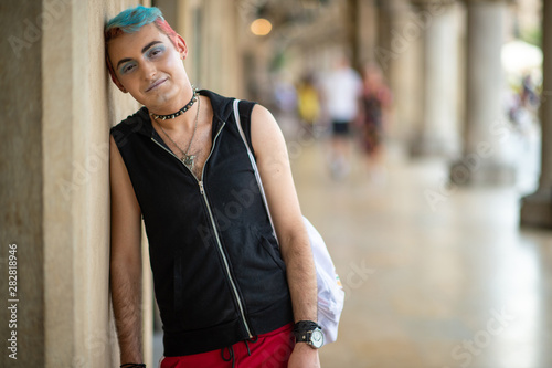 Transgender man in stylish clothing with makeup and colored hairstyle lifestyle portrait in the city with people on background.