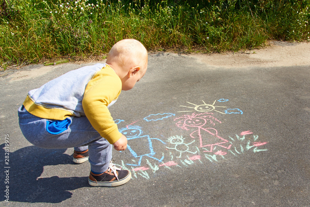Naklejka premium Little boy drawing with chalk on the asphalt, happy family: dad, mom and baby outdoors