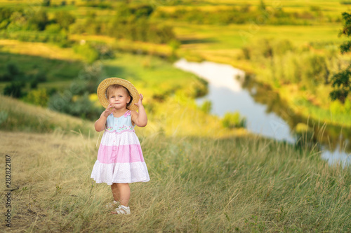 Authentic baby girl in straw hat and cute dress on the walk. Summer meadow. River.