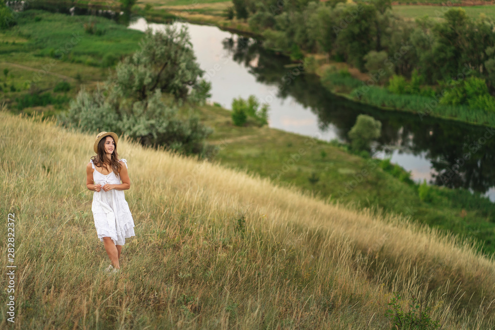 Beautiful girl in a straw hat and a white dress on a walk near the river
