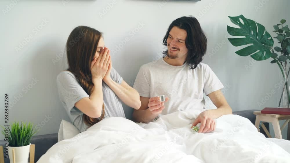 Young man is making marriage proposal to surprised girl putting ring on finger in bed at home laughing kissing hugging. Family, love and relationship concept. Young man is making marriage proposal to surprised girl putting ring on finger in bed at home laughing kissing hugging. Family, love and relationship concept.