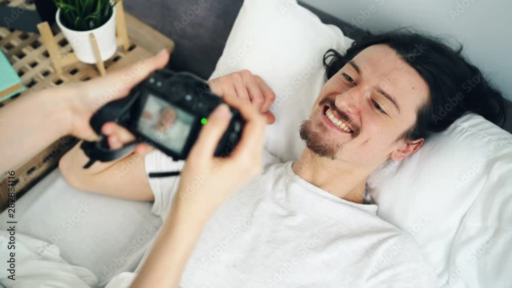 Cheerful male student posing for camera at home in bed laughing having fun while long-haired girlfriend taking pictures. People, hobby and joy concept. Cheerful male student posing for camera at home in bed laughing having fun while long-haired girlfriend taking pictures. People, hobby and joy concept.