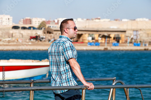 Portrait of tourist and traveler is posing and walking on beach pier, enjoys in sun and view of sea landscape. Man with sunglasses on vacation in summertime in hotel resort on sea jetty platform.