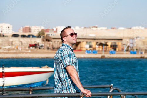 Portrait of tourist and traveler is posing and walking on beach pier, enjoys in sun and view of sea landscape. Man with sunglasses on vacation in summertime in hotel resort on sea jetty platform.