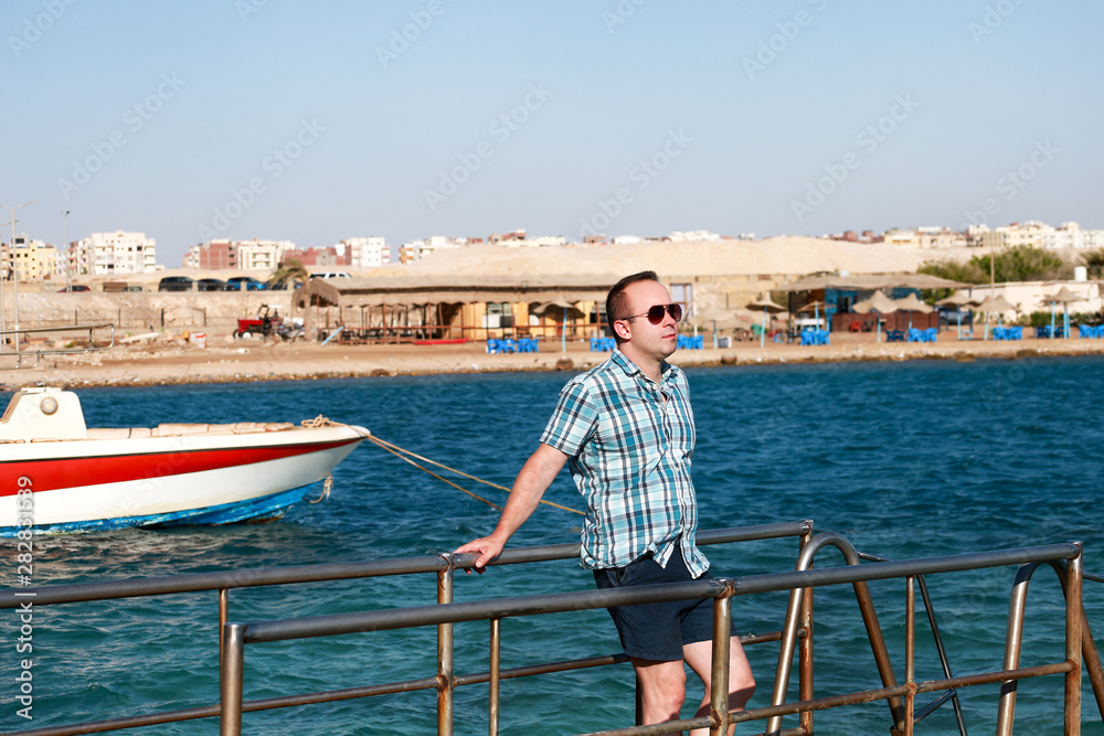 Portrait of tourist and traveler is posing and walking on beach pier, enjoys in sun and view of sea landscape. Man with sunglasses on vacation in summertime in hotel resort on sea jetty platform. Portrait of tourist and traveler is posing and walking on beach pier, enjoys in sun and view of sea landscape. Man with sunglasses on vacation in summertime in hotel resort on sea jetty platform.
