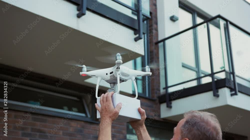 White quadcopter flying in the city with parcel box. Close-up handsome man customer receiving a delivery grabbing the box in hands.