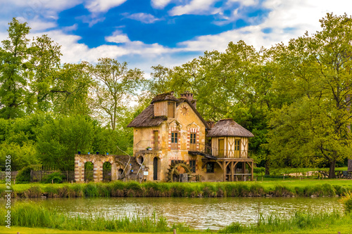 Picturesque view of the Mill, a rustic building in the Queen's Hamlet, surrounded by trees under a blue sky in front of a lake in the park of the Château de Versailles built for Marie Antoinette.