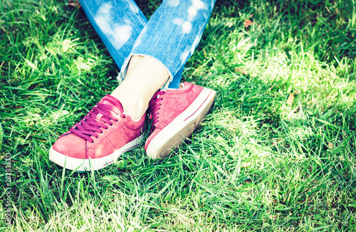 Young woman legs in sport shoes sneakers of pink suede, sitting on the grass lawn in park.