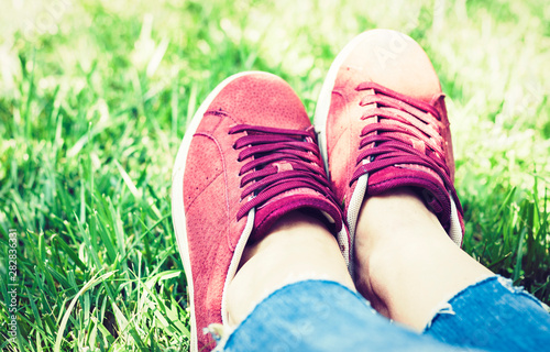 Young woman legs in sport shoes sneakers of pink suede  sitting on the grass lawn in park.
