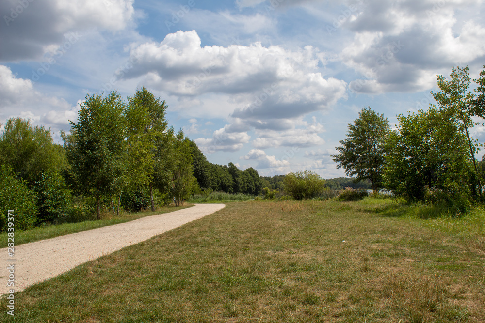 Beautiful summer landscape of a country road in a green meadow surrounded by trees and a lake.