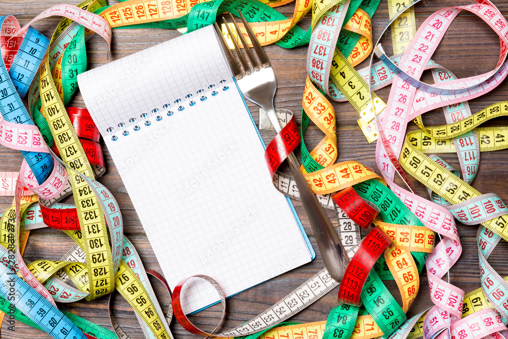 Fototapeta premium Group of colorful measure tapes, open notebook and fork on wooden background with empty space for your idea. Top view of healthy lifestyle concept