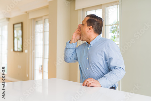 Middle age man sitting at home shouting and screaming loud to side with hand on mouth. Communication concept.