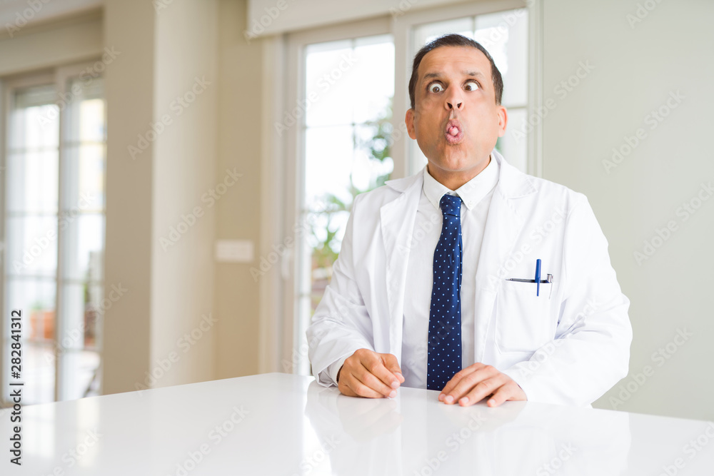 Middle age doctor man wearing medical coat at the clinic making fish face with lips, crazy and comical gesture. Funny expression. Middle age doctor man wearing medical coat at the clinic making fish face with lips, crazy and comical gesture. Funny expression.