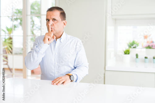 Middle age man sitting at home asking to be quiet with finger on lips. Silence and secret concept.